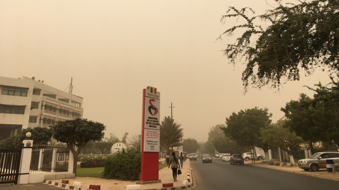 A sign reading "Ministère de la Santé et de l'Action Sociale," with a Senegalese flag, is featured in an image of a Dakar street on a dusty day.